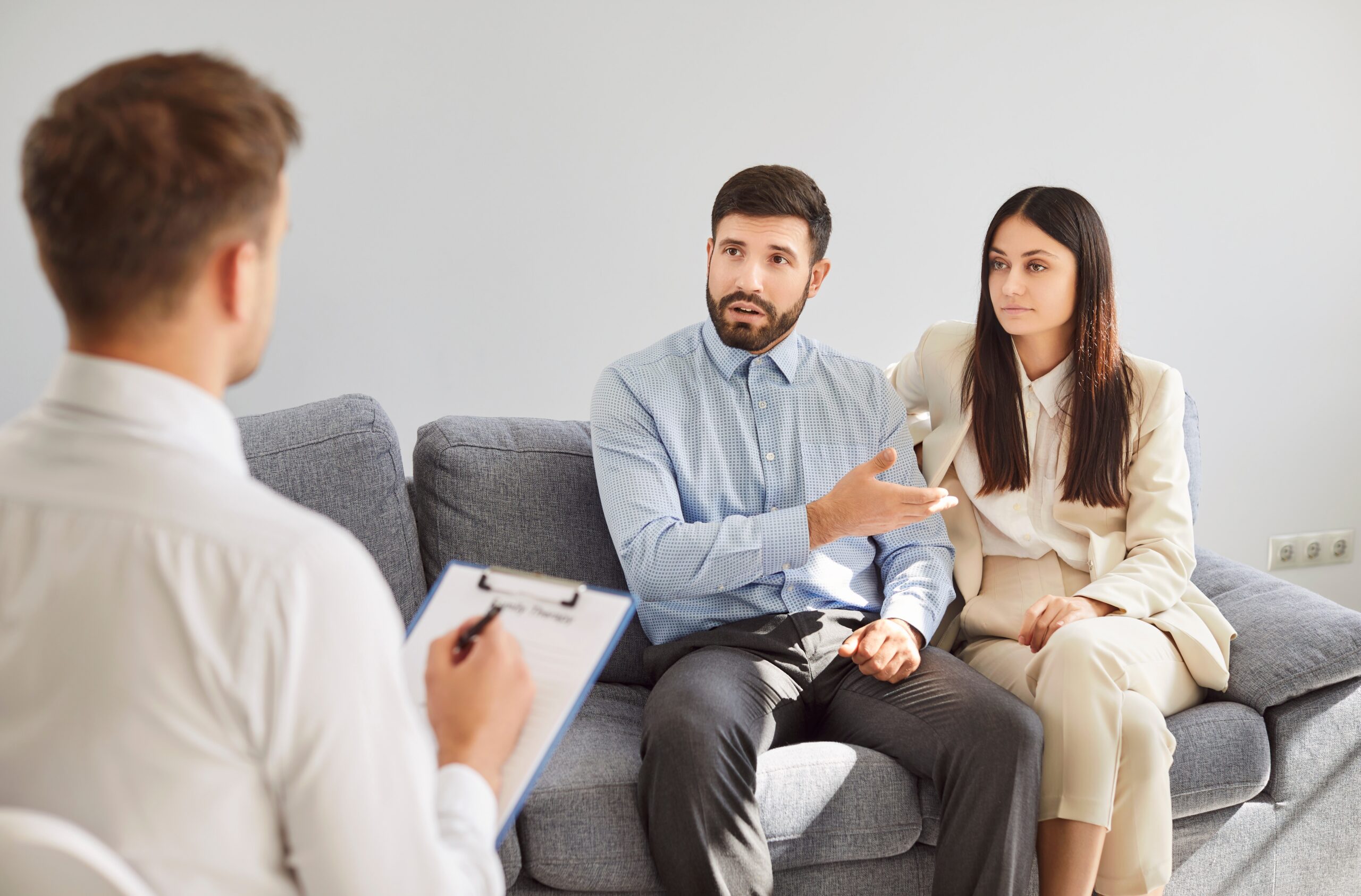 A couple sitting on a couch talking to a therapist holding a clipboard.